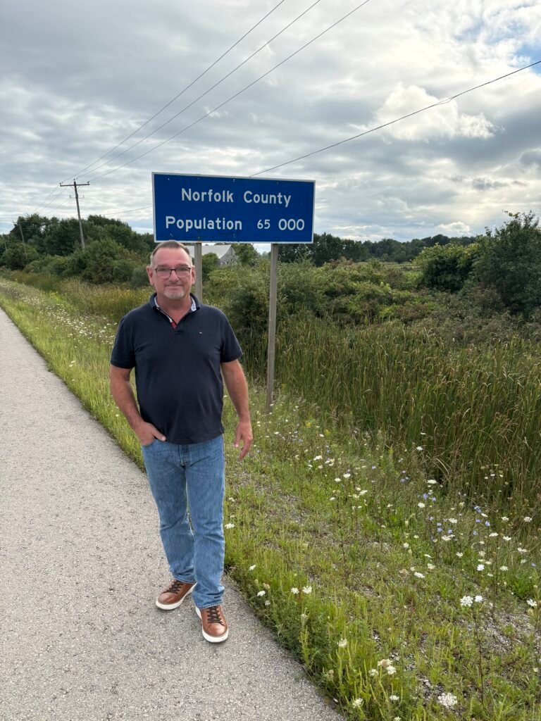 Bill Jukes standing beside the Norfolk County population sign on a rural road